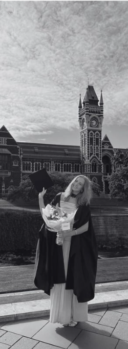 Elvie Fox in graduation regalia holding flowers, in front of a historic building with a clock tower